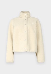 Kermanvärinen fleece-takki, jossa on korkea kaulus, nepparit ja lyhyt leikkaus. Pehmeä rakenne, jossa on minimaalinen brändäys.