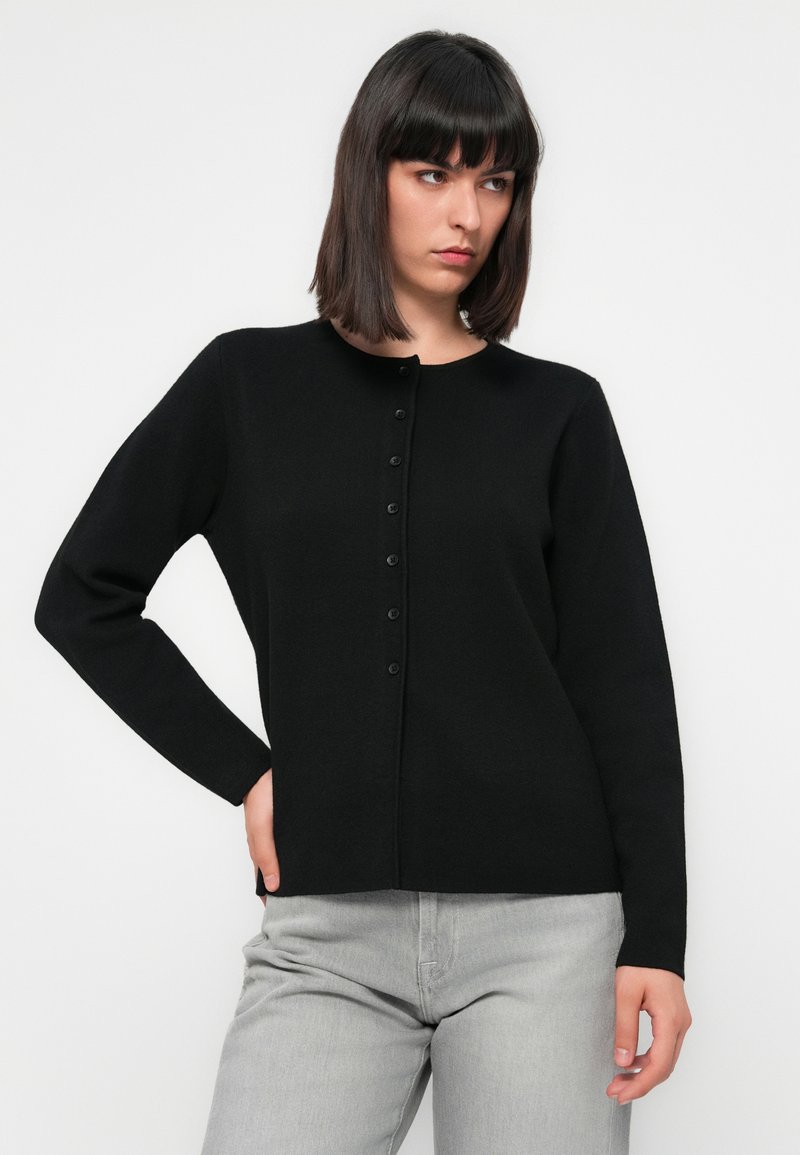 Czarny kardigan zapinany na guziki z długimi rękawami, wykonany z miękkiego materiału. Posiada okrągły dekolt i subtelną teksturę. Noszony z jasnoszarymi spodniami.