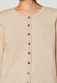 Light beige cardigan made of soft fabric. Features a rounded neckline and a front button closure with six dark buttons along the placket.