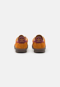 Baskets en suède orange avec un accent bordeaux au talon, dotées d'une semelle en caoutchouc et d'un logo en relief à l'arrière.