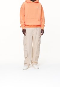 Oranžová mikina s kapucí a přední kapsou, doplněná béžovými cargo kalhotami s několika postranními kapsami a bílými teniskami.