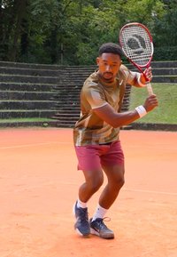Tennis spiller i en brunt og grønt mønstret skjorte og bordeaux shorts, som holder en racket, på en oransje leirbane med gressbakgrunn.