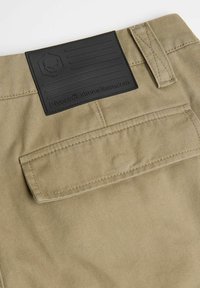 Spodnie z khaki materiału z teksturowaną czarną gumową metką z wzorem czaszki i kieszenią z tyłu. Widoczne są przeszywane akcenty.