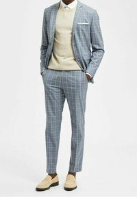 Ljust blårandig kostym med avsmalnande byxor, vit skjorta, beige tröja och krämfärgade loafers. Anmärkningsvärd fickdetalj på jackan.