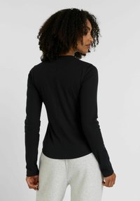Schwarzes Langarmoberteil mit figurnahem Design und gerippter Textur, das einen runden Ausschnitt und nahtlose Rückendetails aufweist. Getragen mit grauen Jogginghosen.