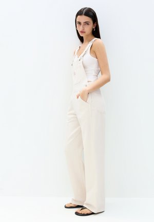 Witte denim overall met wijde pijpen, verstelbare banden, voorzakken en een achterpatch, gedragen over een aansluitend wit topje en gecombineerd met zwarte sandalen.