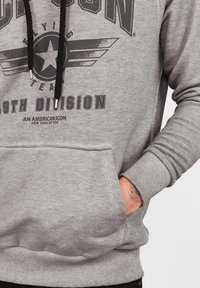 Grå sweatshirt med huva, tryckt logotyp och vingar, framficka, ribbade muddar och justerbara snören. Mjuk, texturerad tyg.