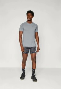 Athletic man standing, wearing a gray Nike T-shirt, black camo-patterned shorts, black Nike socks, and black running shoes against a plain background.