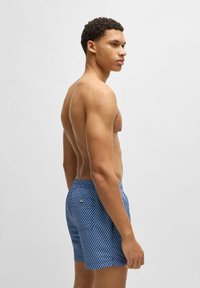 Blue patterned swim shorts with a textured mesh design. Elastic waistband with a small brand tag at the back. Fitted style.