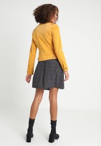 Žena s kudrnatými vlasy, oblečená do hořčicově žluté bundy, černé vzorované sukně a černých kotníkových bot, stojí a dívá se do strany.