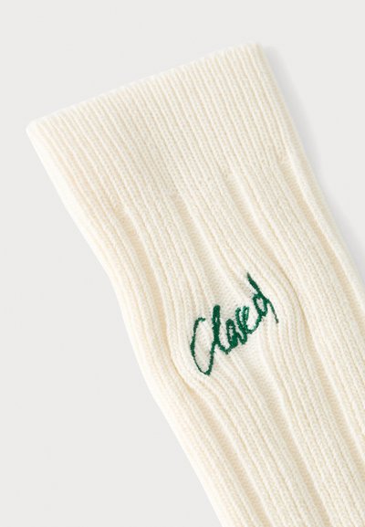 Gros plan d'une chaussette tricotée crème côtelée avec un texte vert brodé au niveau de la cheville.