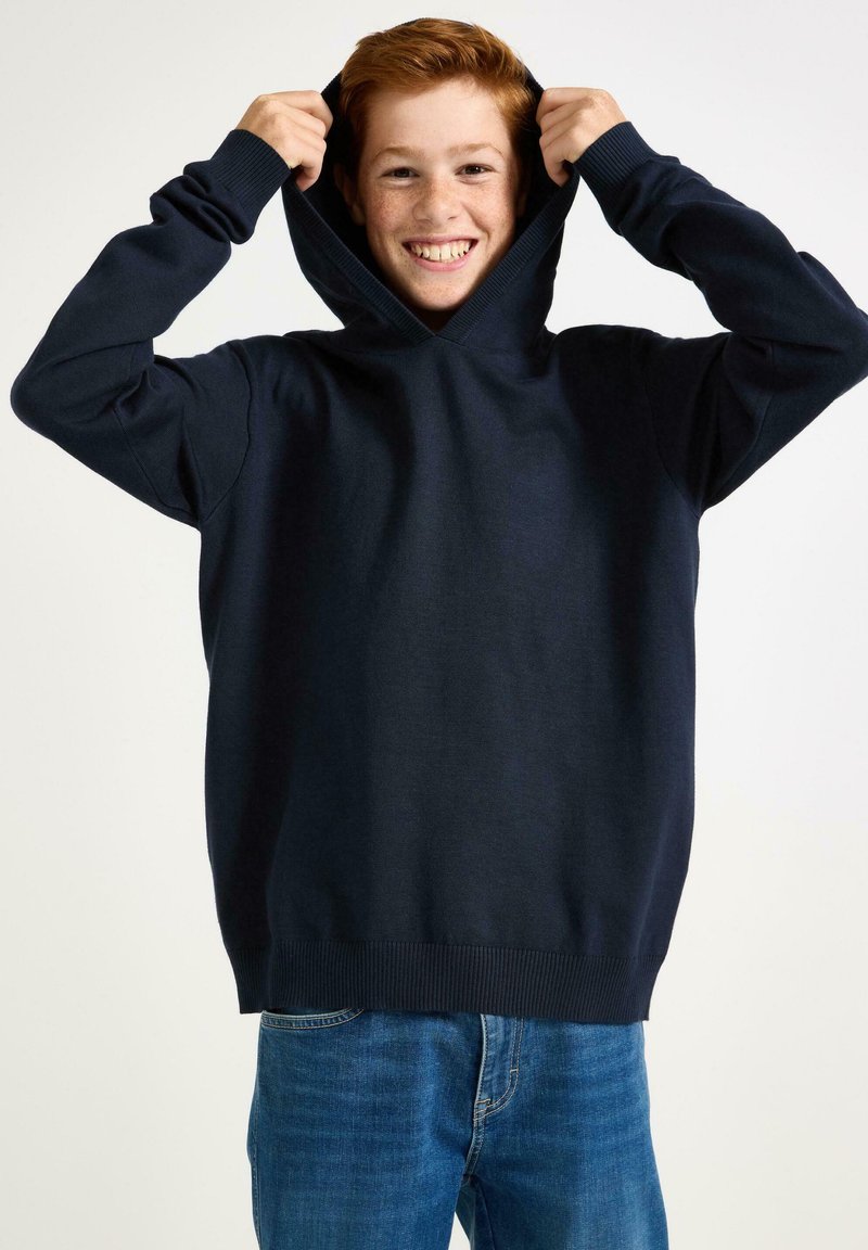 Felpa con cappuccio blu navy, con polsini e orlo a costine. Tessuto morbido, vestibilità comoda e cordino regolabile sul cappuccio. Indossata da un giovane sorridente.