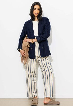 Femme portant un blazer bleu marine, une chemise blanche, un pantalon beige à larges rayures, tenant un sac beige à franges, et portant des chaussures beiges sans lacets.