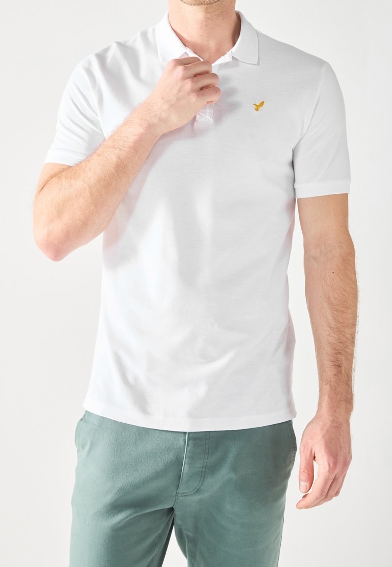 Homme portant un polo à manches courtes blanc avec un petit logo d'oiseau jaune sur la poitrine et un pantalon vert, ajustant le col avec la main.