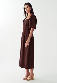 Rjava, srednje dolga obleka s kvadratnim izrezom, napihnjenimi rokavi in tekočim krilom. Nosi se s svetlo bež natikači na blok peto z odprtim prstom.