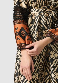 Långärmad klänning i mönstrad svart, beige och orange tyg. Texturerad spets på ärmarna ger detalj. Elastiskt manschettdesign.