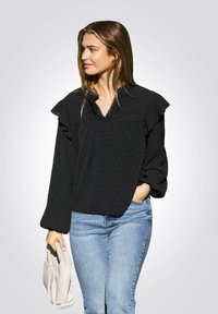 Zwarte blouse met gerimpelde schouders, gestructureerd patroon, kantafwerking bij de halslijn, gecombineerd met blauwe spijkerbroek en een lichtgekleurde tas.