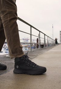 Bottines noires en matériau synthétique avec une finition texturée, dotées d'un système de laçage et d'un accent logo, présentées sur un chemin en béton.