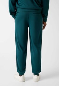 Turquoise joggingbroek gemaakt van gladde stof, met een elastische tailleband, geribbelde boorden en een achterzak. Gecombineerd met witte sneakers.