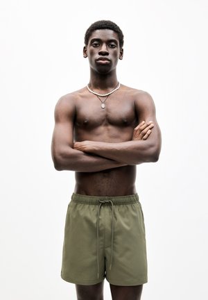 Short olive green swim shorts with an elastic waistband and drawstring. The model has a muscular build and wears silver accessories.