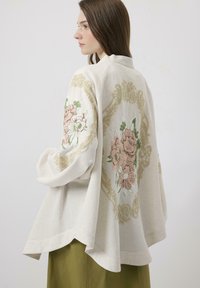 Jachetă brodată de culoare crem, cu desene florale în nuanțe de roz și verde pe mâneci; croială lejeră cu margine rotundă și textură moale.