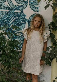 Kermanvärinen mekko, jossa on teksturoitu kuvio, ja rypytetyt hihat, joissa on sinisiä kukkaistutuksia. Ympäröivät vihreät lehdet, jotka ovat taustalla muurissa.