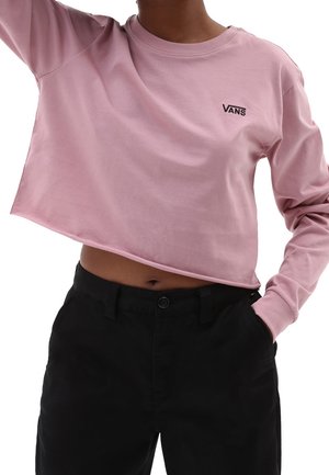 Osoba w różowej, przyciętej bluzie Vans z długim rękawem, w połączeniu z czarnymi spodniami z wysokim stanem, jedną ręką w kieszeni, na białym tle.