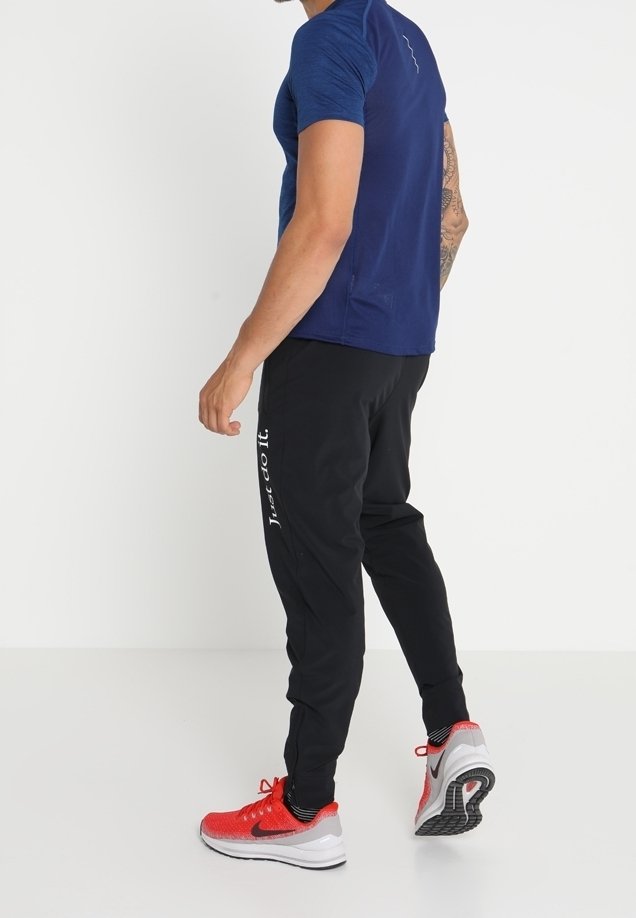 Muž v modré košili s krátkým rukávem, černých joggingových kalhotách s textem "Just do it." a červených teniskách Nike stojí proti bílému pozadí.
