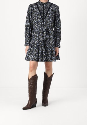 Kobieta ubrana w czarną kwiatową sukienkę z długimi rękawami i paskiem w talii, do tego brązowe kowbojskie buty z wysoką cholewką sięgającą do kolan.