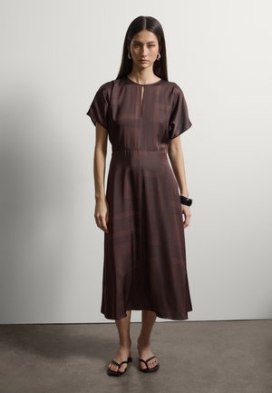 Femme aux longs cheveux bruns portant une robe midi marron à motifs géométriques, manches courtes, bracelet noir et tongs noires, debout contre un mur uni.