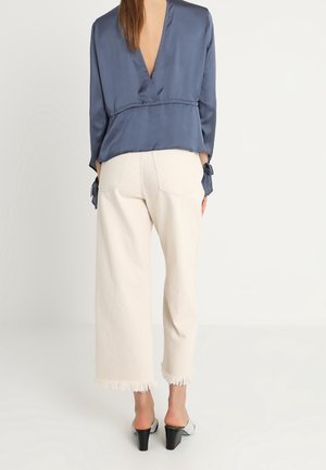 Kvinde iført en blå bluse med lange ærmer og dyb V-udskæring bagpå samt hvide cropped jeans med frynser og sorte mulehøjhælede sko, stående mod en hvid baggrund.