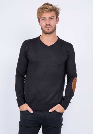 Jeune homme aux cheveux blonds portant un pull noir à col en V avec des coudières marron, debout les mains dans les poches devant un fond uni.