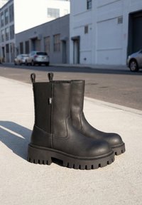 Schwarze Leder-Stiefeletten mit klobiger Sohle und elastischen Seitenteilen, die bei Tageslicht auf einem Bürgersteig in der Stadt stehen.