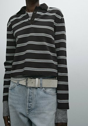 Personne portant une chemise à manches longues rayée noir et blanc sur des manches grises, un jean bleu clair et une ceinture texturée de couleur claire.
