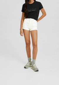 Camiseta negra con texto impreso en blanco, shorts altos blancos, calcetines grises y zapatillas grises con estampado. Diseño minimalista, ajuste casual.