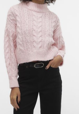 Woman wearing a light pink chunky cable-knit sweater and high-waisted black pants with a black belt, hand in pocket.