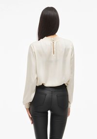 Blusa de seda crema con mangas largas, cuello smock y cierre de ojal con botón en la parte posterior, combinada con pantalones negros ajustados.