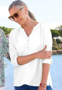 Weiße Bluse mit V-Ausschnitt und aufgerollten Ärmeln. Glatte Textur, lässiges Design, kombiniert mit einem roten Armband. Im Hintergrund sind Wasser und Grün zu sehen.