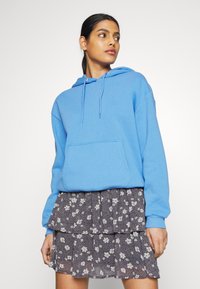 Camisola azul com cordões ajustáveis e bolso frontal, sobreposta a uma saia escura com padrão floral e Babados. Textura de tecido suave.