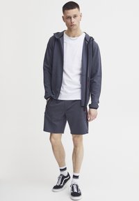 Sivý zip-up mikina s čiernymi akcentmi, biele tričko, sivé šortky, čierne tenisky s bielymi podrážkami a pruhované ponožky. Neformálny športový outfit.