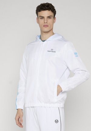 Junger Mann trägt einen weißen Sergio Tacchini Reißverschluss-Hoodie mit hellblauen Streifen an den Ärmeln und passende weiße Hosen mit Logodetails.