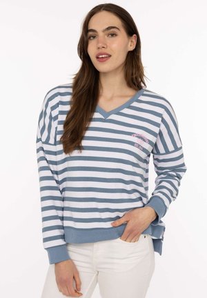 Junge Frau trägt einen blau-weiß gestreiften V-Ausschnitt-Pullover mit Text und Herzdetail, kombiniert mit weißen Hosen, posiert vor weißem Hintergrund.