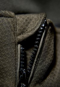 Mörk olivfärgad tyg med en mjuk, texturerad yta. Har en svart dragkedja med synliga tänder och en något upphöjd söm vid kanten.