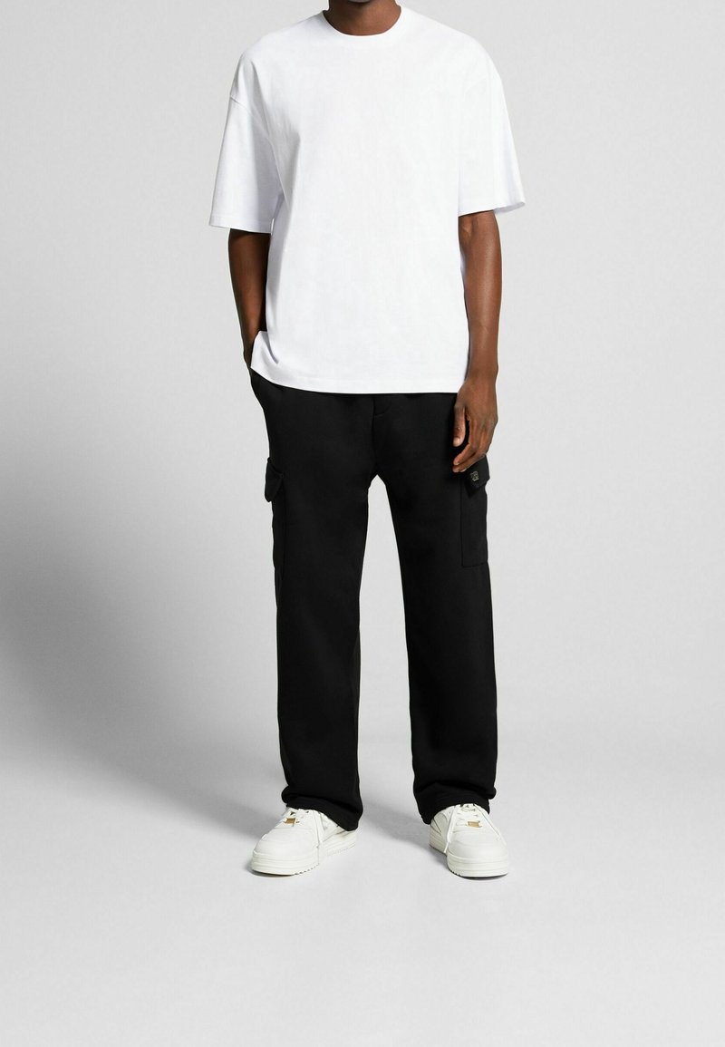 Homme portant un t-shirt oversize blanc uni, un pantalon cargo noir avec poches, et des baskets blanches, debout devant un fond gris clair.