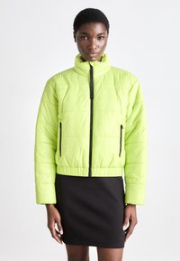 Veste rembourrée verte fluo avec un col haut, fermeture éclair noire et poches latérales. Présente une texture matelassée et une taille élastique.