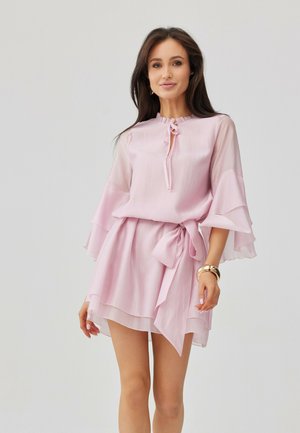 Kobieta ubrana w jasnoróżową, warstwową sukienkę mini z rozkloszowanymi rękawami i paskiem w talii, stojąca na jednolitym szarym tle.