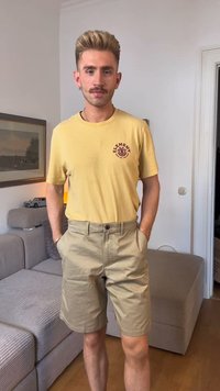 Žuta majica s kratkim rukavima s bordo logotipom, u kombinaciji s svijetlim kaki šortsevima. Model stoji s rukama u džepovima, ispred neutralne pozadine.