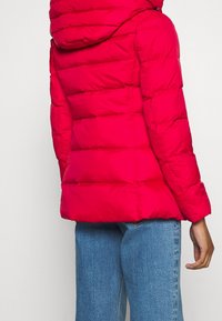Doudoune rouge avec capuche, présentant des sections matelassées horizontales, une fermeture éclair à l'avant et des poignets élastiques, associée à un jean bleu.