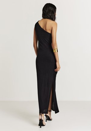 Woman wearing a black one-shoulder maxi dress with a side slit, black high-heeled sandals, and a gold arm cuff, standing against a plain background.