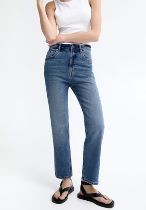 Femme portant un jean bleu taille haute, coupe droite, un débardeur blanc et des sandales plates noires, debout devant un fond blanc.
